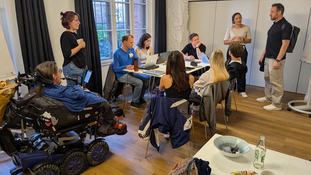 Das Foto zeigt eine Gruppe von Menschen, die an einem Tisch sitzen. Eine Person sitzt in einem besonderen Roll-Stuhl.