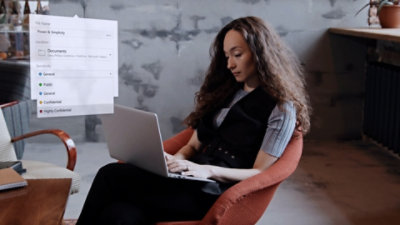 A woman sitting on a chair and working on a laptop keeping on her lap and some window open.