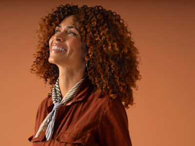 A person with curly hair, wearing a rust-colored jacket and a striped scarf, looks up with a smile