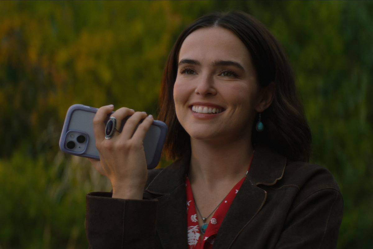 A smiling woman with dark hair holds a smartphone outdoors, wearing a red patterned shirt, a brown jacket, and turquoise earrings, with blurred green foliage in the background.