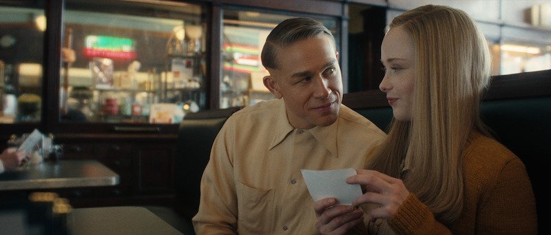 A man and a woman sit closely together in a diner booth, smiling warmly at each other while the woman holds a small piece of paper.