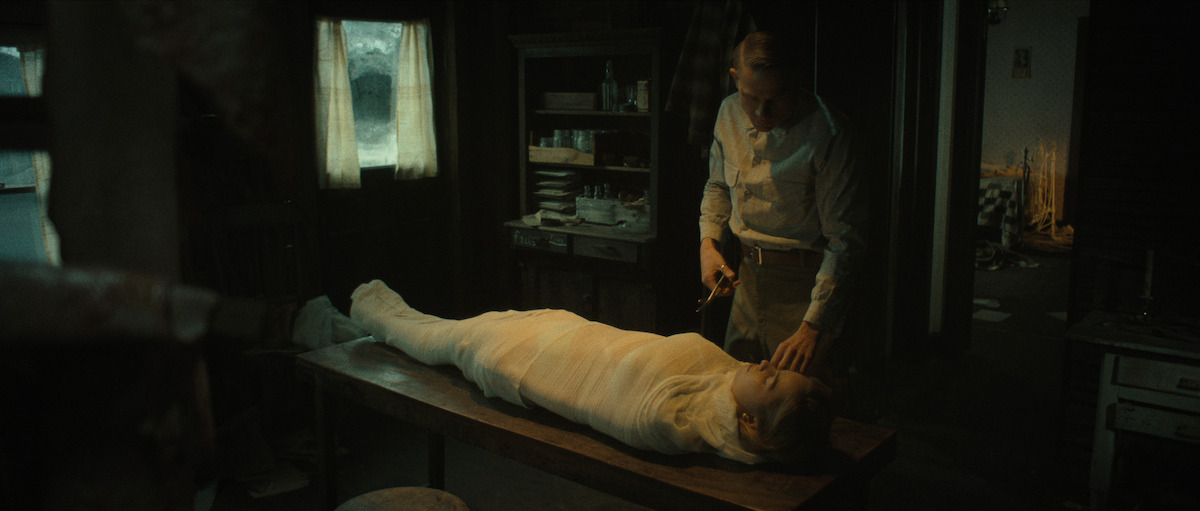 A person wrapped entirely in white bandages lies on a table while another person stands beside them holding scissors in a dimly lit room.