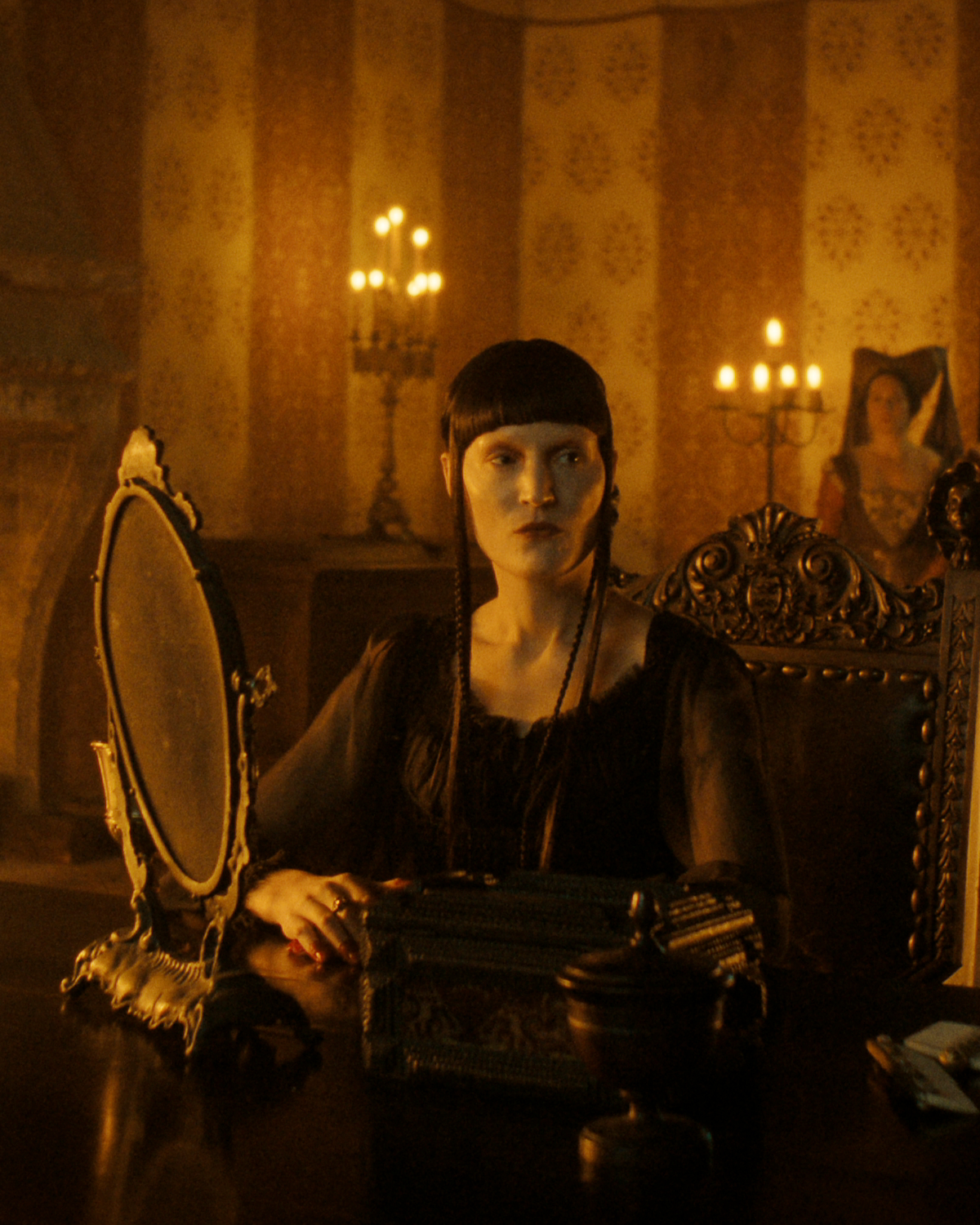 A woman in dark, vintage clothing sits at a lavish wooden vanity with ornate mirror, lit by warm candlelight in an opulent, richly decorated room with patterned wallpaper and antique furnishings.