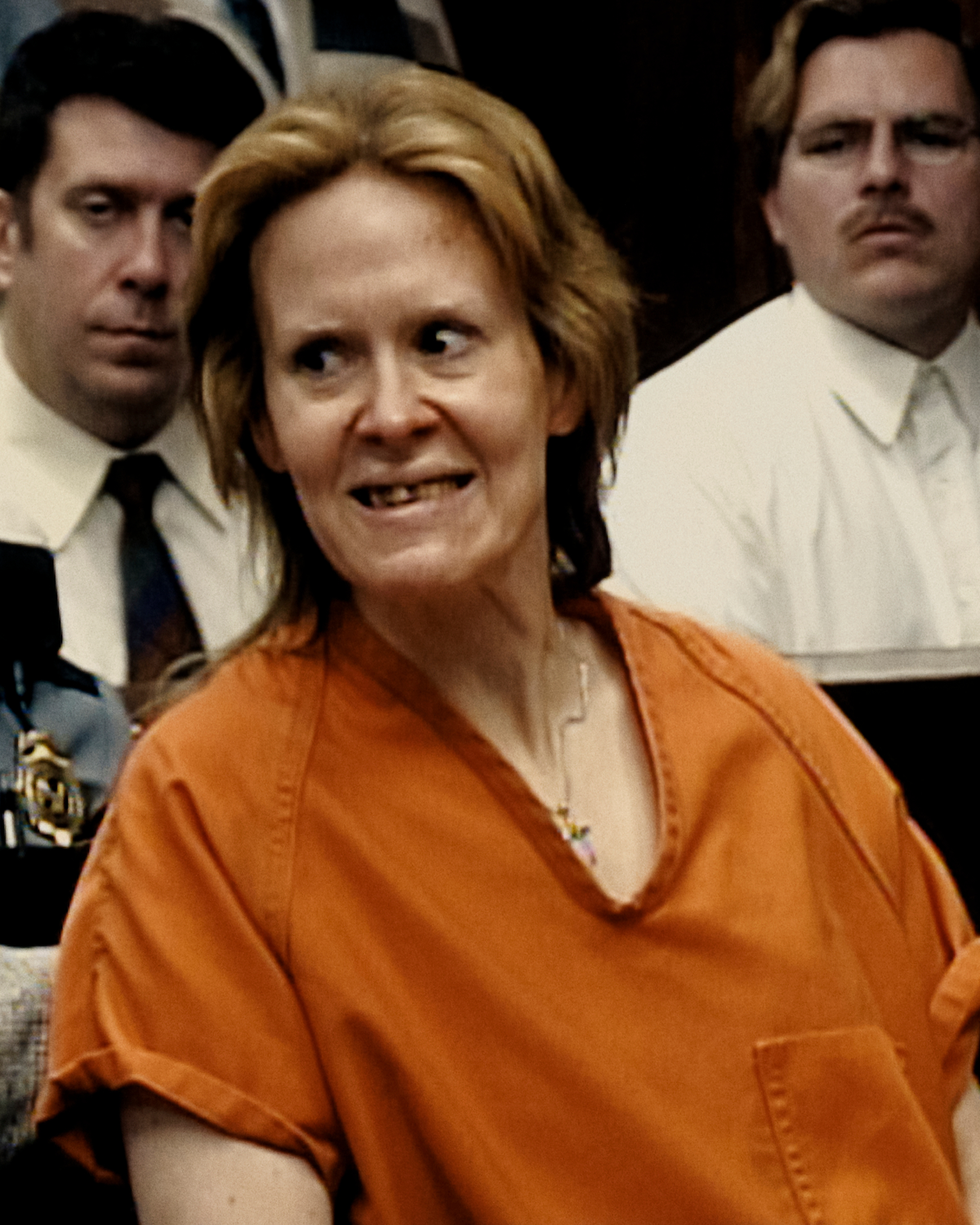 Woman in orange prison uniform sits in a courtroom, surrounded by people in formal attire, under indoor lighting, with focused and serious expressions on faces in the background.