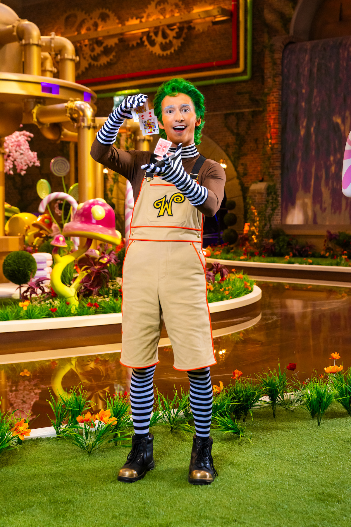 Person dressed as an Oompa Loompa with green hair and striped clothing stands on a whimsical, candy-themed set with colorful plants, large pipes, and fantasy decorations, holding a chocolate bar with a happy expression.