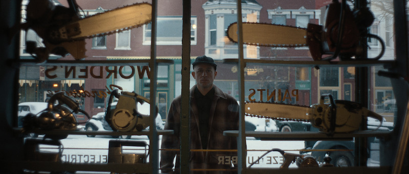 A man in a cap and plaid jacket stands outside a hardware store window, looking in past shelves filled with chainsaws and tools on a snowy day.