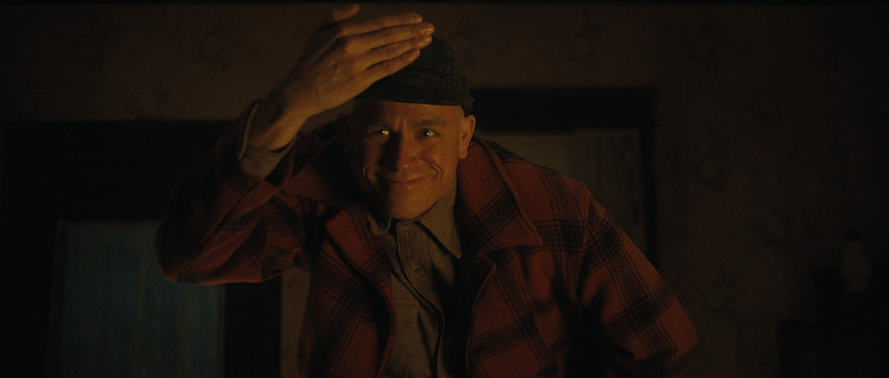 A man in a plaid jacket and cap smiles warmly while tipping his hat in a dimly lit room.