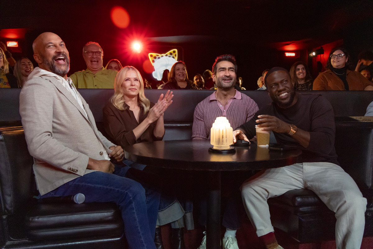 Four people sit at a round table in a dimly lit comedy club, laughing and clapping. An audience is visible in the background, also smiling and enjoying the lively atmosphere. A candle sits on the table.