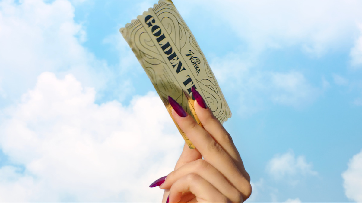 A hand with long, painted nails holds up a golden ticket against a blue sky with clouds.