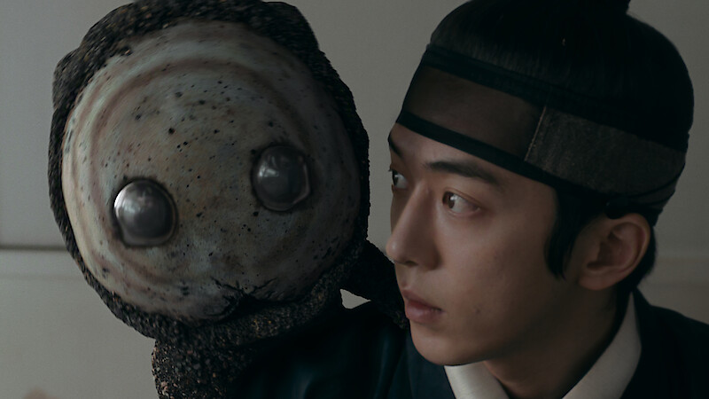 A young man in traditional Korean attire closely observes a large, round, alien-like puppet or creature with big metallic eyes in an indoor setting, suggesting a scene from a historical drama with fantasy elements.