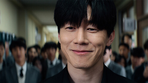 A young man with black hair smiles confidently in a school hallway, surrounded by other students in uniforms, with bright lighting and a lively environment.