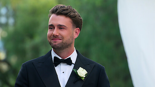 A man in a black tuxedo with a white rose boutonniere stands outside, smiling with greenery in the background, likely at a formal event such as a wedding.