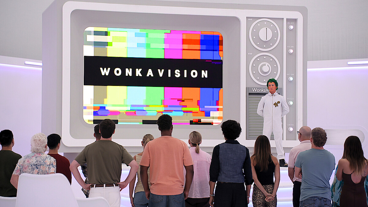 A group of people watch a large retro television screen displaying “WONKAVISION” with colorful test patterns, in a futuristic, white room. A person in a white lab coat stands beside the TV, leading the presentation.