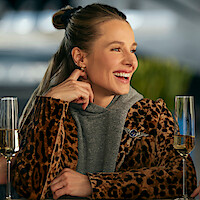 Smiling woman in a leopard print jacket and gray hoodie sitting at a table with two glasses of champagne, in a modern indoor setting with soft lighting and blurred green plants in the background.