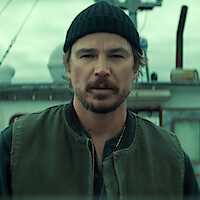 A man in a knit cap and vest stands on a boat with a weathered exterior, cloudy sky in the background, creating a rugged, maritime atmosphere.