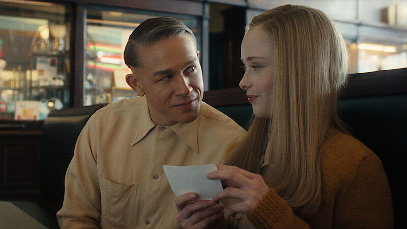 A man and woman, seated in a diner booth, share a sweet moment, smiling and holding a piece of paper between them.