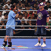 Two wrestlers face off in a crowded wrestling ring, one wearing a blue shirt speaking into a microphone, the other in a purple shirt holding a championship belt. The audience watches intently from all sides.