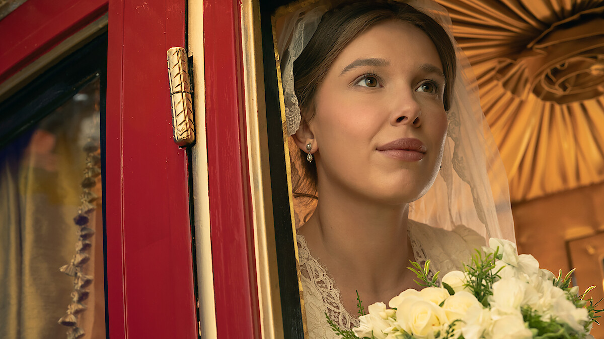 A bride in a wedding dress and veil holding a bouquet of white flowers looks out of a red carriage window with soft lighting and a decorative interior ceiling behind her.
