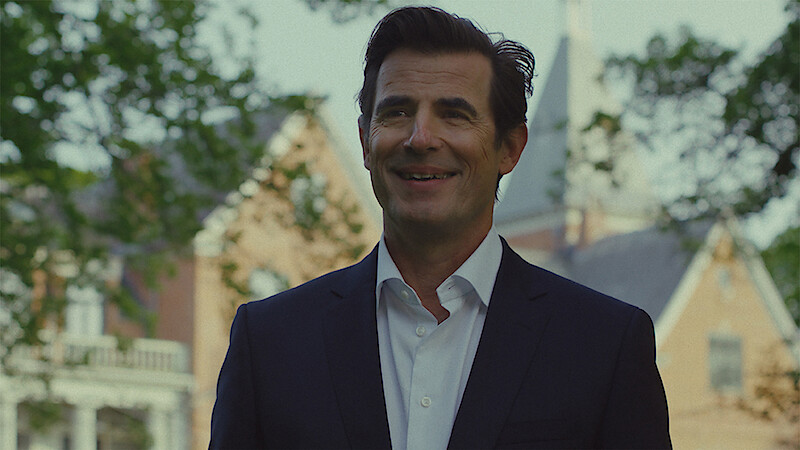 A smiling man in a dark suit stands outdoors in front of a large brick building with trees and a steeple visible in the background, suggesting a formal or academic setting on a sunny day.