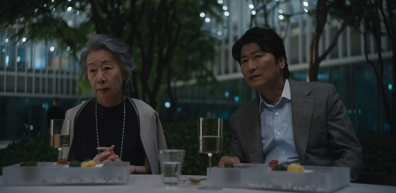 Youn Yuh-jung as Chairwoman Park and Song Kang-ho as Dr. Kim sit at an outdoor table in an urban garden at night, with food, drinks, and modern buildings lit up in the background, creating a sophisticated and calm atmosphere.