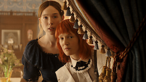 Two women in vintage clothing stand closely together, peering out from behind ornate curtains in an elegant, softly lit room with classic decor and floral arrangements, suggesting a historical or period drama setting.