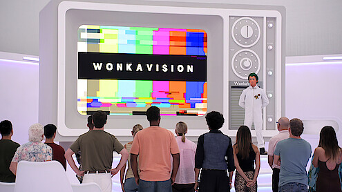 A group of people watch a large retro television screen displaying “WONKAVISION” with colorful test patterns, in a futuristic, white room. A person in a white lab coat stands beside the TV, leading the presentation.