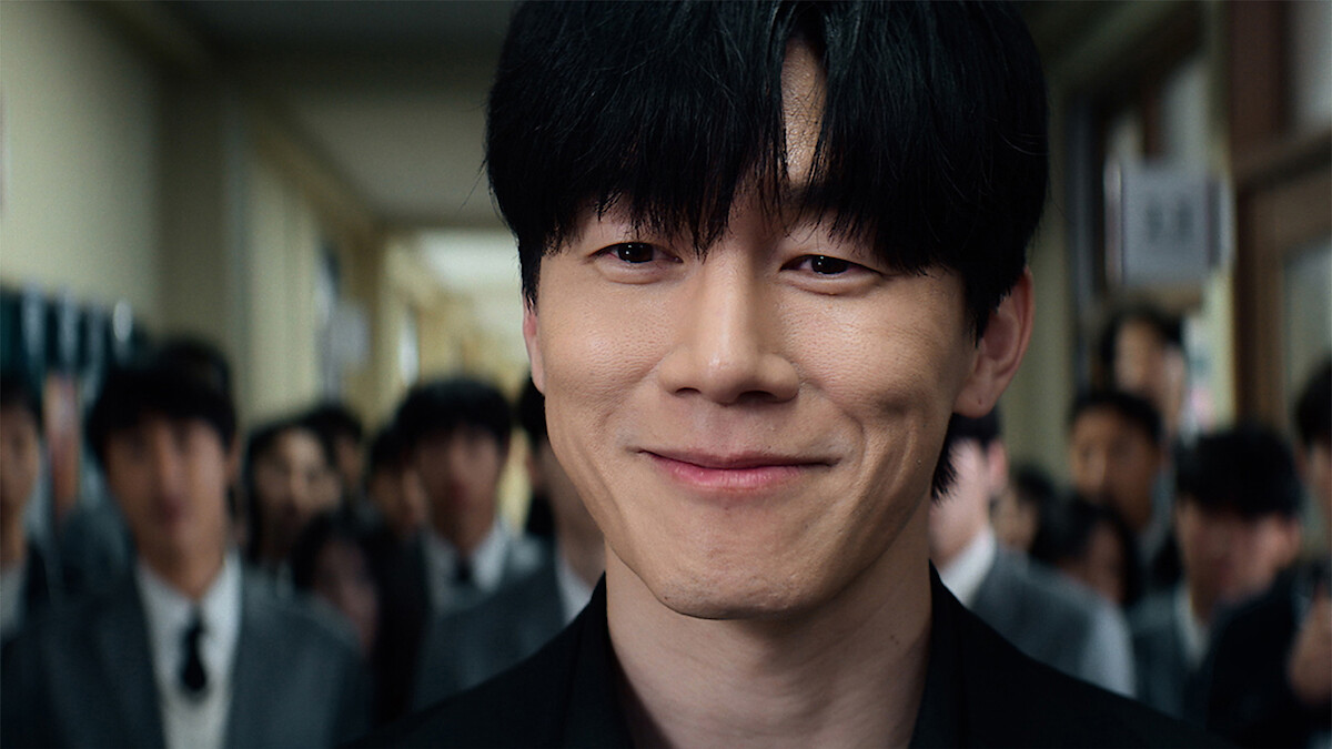 A young man with black hair smiles confidently in a school hallway, surrounded by other students in uniforms, with bright lighting and a lively environment.
