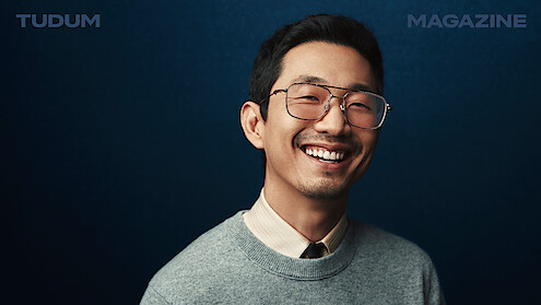 Lee Sung Jin stands in front of a backdrop that shifts from black to dark blue, smiling and looking to camera.