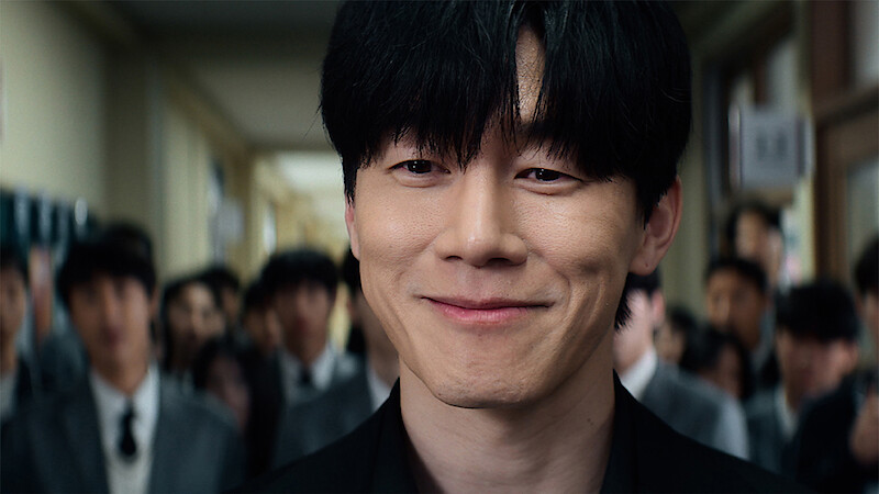 A young man with black hair smiles confidently in a school hallway, surrounded by other students in uniforms, with bright lighting and a lively environment.