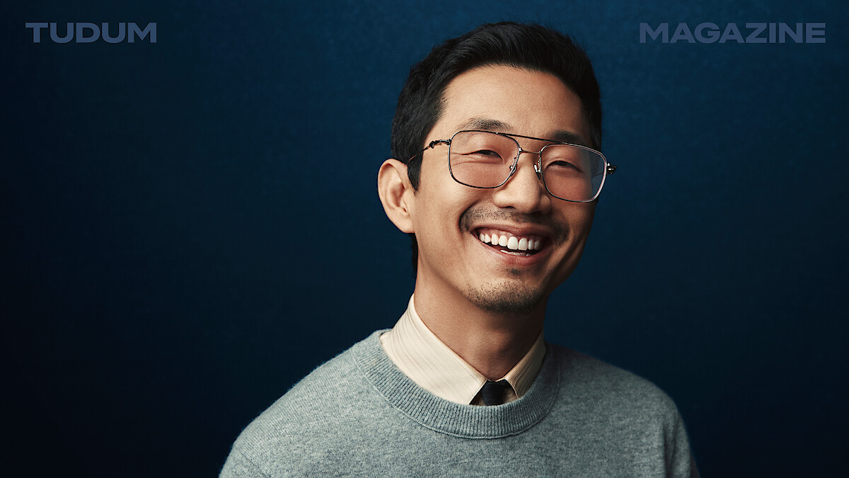 Lee Sung Jin stands in front of a backdrop that shifts from black to dark blue, smiling and looking to camera.