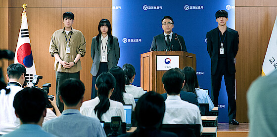 A group of four people face a group of reporters at a press conference.