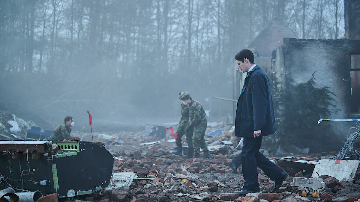 A man in a suit walks through a debris-strewn, foggy ruin as two people in camouflage uniforms investigate the wreckage; trees and a partially destroyed building are in the background.