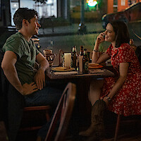 A man and woman sit at a table in a cozy restaurant at night, smiling and talking over drinks and food, creating a warm, intimate atmosphere.