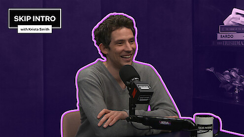 Josh O'Connor smiles while speaking into a podcast microphone in a purple studio, outlined with a neon accent, with a “Skip Intro with Krista Smith” graphic in the corner.