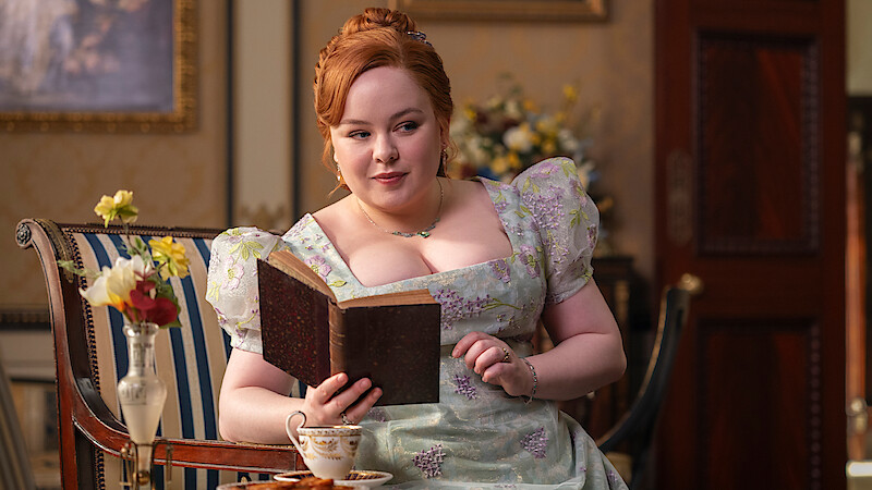 A woman in a floral dress sits in an elegant room with classic decor, reading a book and holding a teacup. Flowers and ornate furniture are in the background, evoking a historical or period setting.