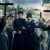 A woman in Victorian-era clothing, handcuffed and escorted by a police officer, stands amid an angry protest crowd holding signs and torches outdoors, suggesting a historical or dramatic arrest scene.