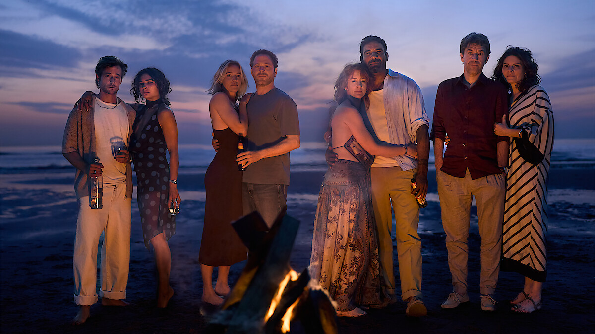 Four couples stand together on a beach at sunset near a campfire, dressed in casual evening clothes, some holding drinks, ocean and colorful sky in the background.