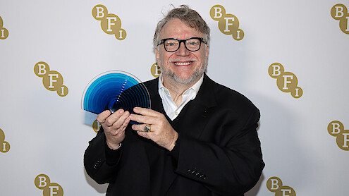 Smiling man in glasses and a suit holding a blue translucent award, standing in front of a backdrop with repeated BFI logos at an indoor event.