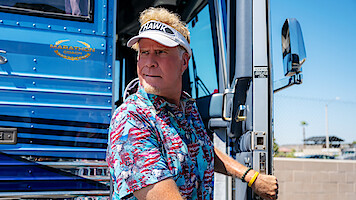 Man in a tropical shirt and visor stands at the doorway of a blue luxury bus on a sunny day, holding the door handle, with a fence and clear sky in the background.