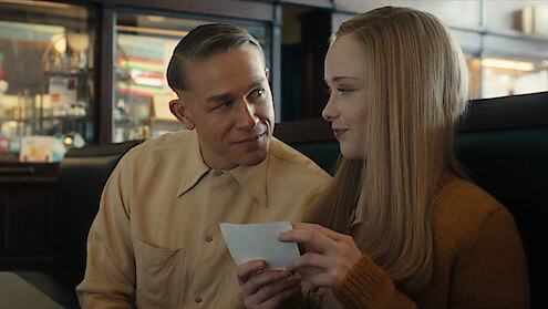 A man and woman, seated in a diner booth, share a sweet moment, smiling and holding a piece of paper between them.