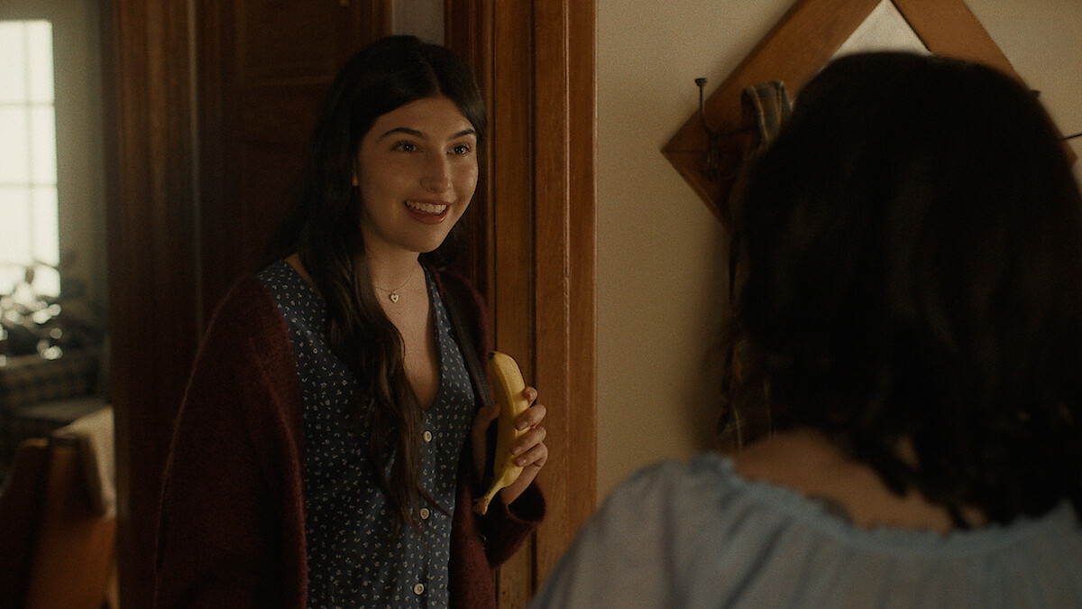 A young woman holding a banana smiles at another person in a cozy, warmly lit room with wooden doors and a wall mirror, suggesting a casual home conversation.