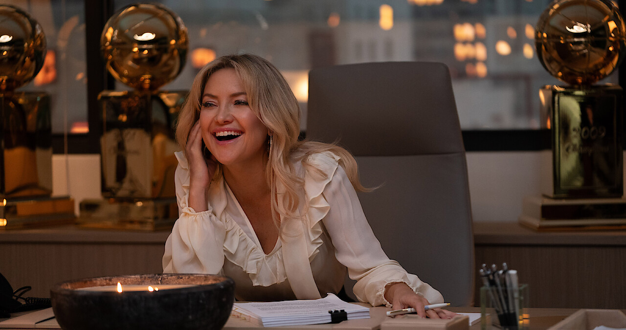 Kate Hudson as Isla Gordon sits at a desk smiling surrounded by basketball trophies in season 1 of 'Running Point'
