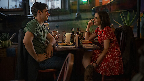A man and woman sit across from each other at a cozy restaurant table at night, smiling and talking over food and drinks, with a street visible through the window behind them.