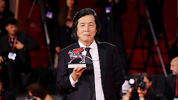 Man in a suit holding a silver lion trophy on a red carpet, surrounded by photographers and people with cameras and badges in an event or awards ceremony setting.