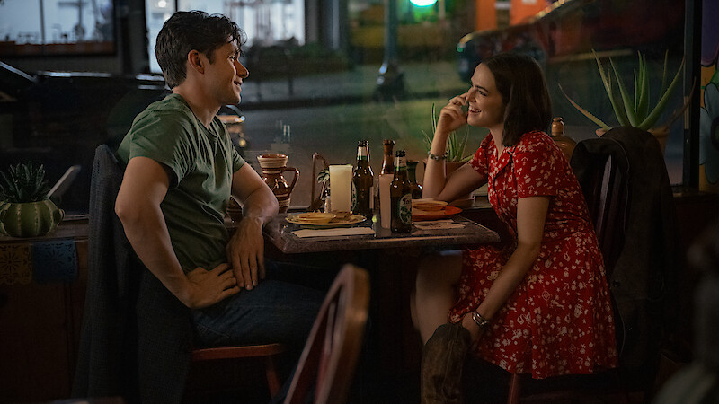 A man and woman sit across from each other at a cozy restaurant table at night, smiling and talking over food and drinks, with a street visible through the window behind them.