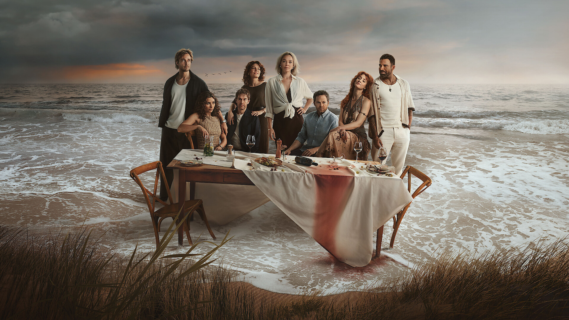 Eight people pose around a table with food and drinks on a beach at sunset, with dramatic waves and clouds behind them. The tablecloth is stained red, suggesting a mysterious or dramatic atmosphere.
