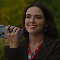 Smiling woman with dark hair holding a phone outdoors, wearing a brown jacket, red shirt, turquoise jewelry, and standing in front of a blurred green background.
