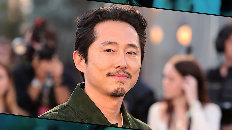 Steven Yeun, com cabelo escuro curto e barba aparada, veste uma jaqueta verde. Ele está ao ar livre em um evento com pessoas, câmeras e luzes desfocadas ao fundo, criando um clima profissional e animado.