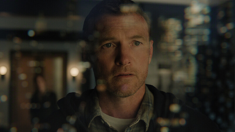 A man stands indoors, looking intently through a glass wall with light reflections; the environment is dimly lit with modern decor, and a blurred figure is visible in the background.