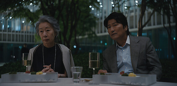 Youn Yuh-jung as Chairwoman Park and Song Kang-ho as Dr. Kim sit at an outdoor table in an urban garden at night, with food, drinks, and modern buildings lit up in the background, creating a sophisticated and calm atmosphere.